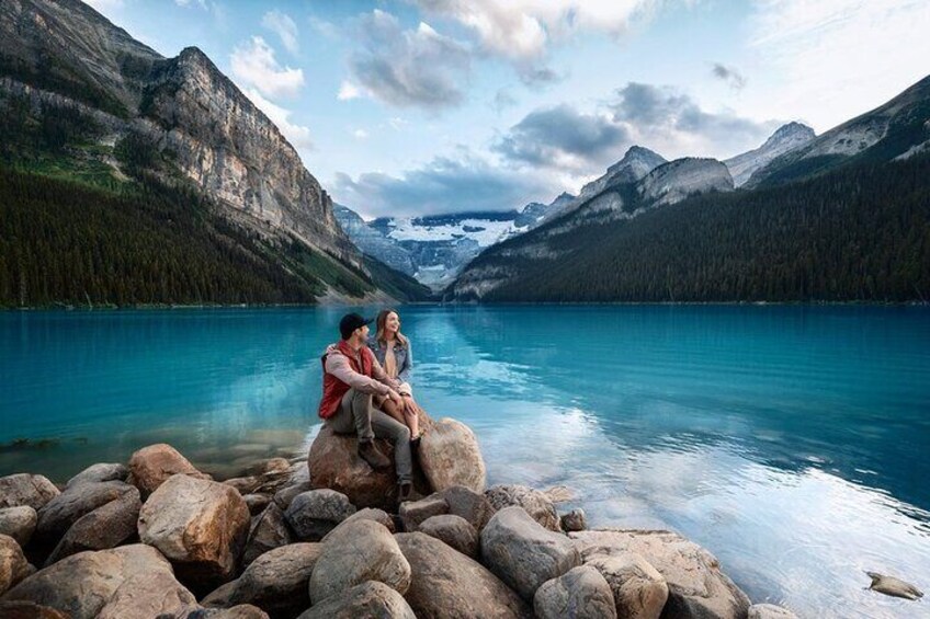 Una pareja sentada sobre rocas junto a un lago, con montañas al fondo. 1