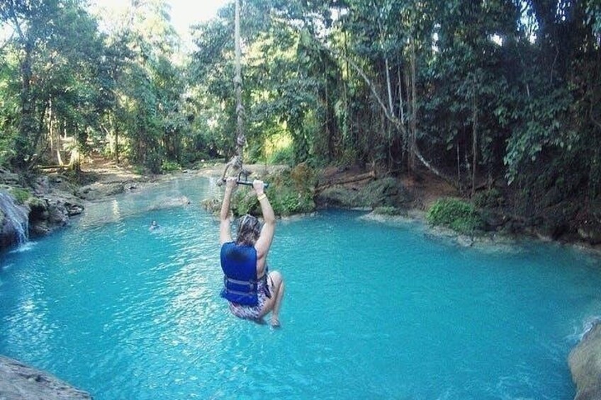 A person in a blue swimsuit hanging from a rope over a clear blue pool surrounded by trees. 2