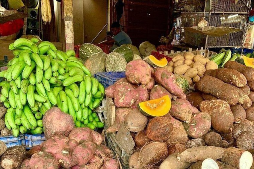 Un puesto de mercado con diversas verduras, entre ellas plátanos verdes, batatas y un calabacín amarillo. 5