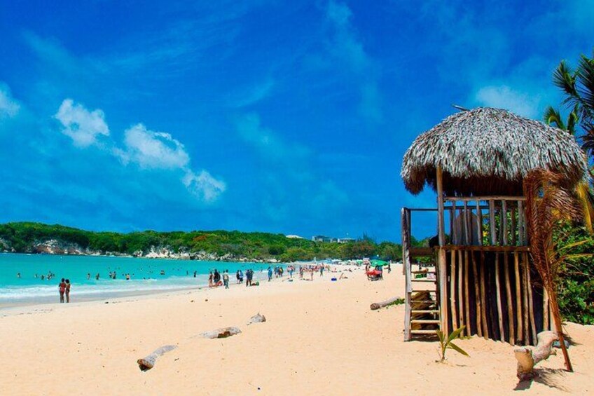 Ein Strand mit einer Reetdachhütte, klarem türkisblauem Wasser und badenden Personen. 5