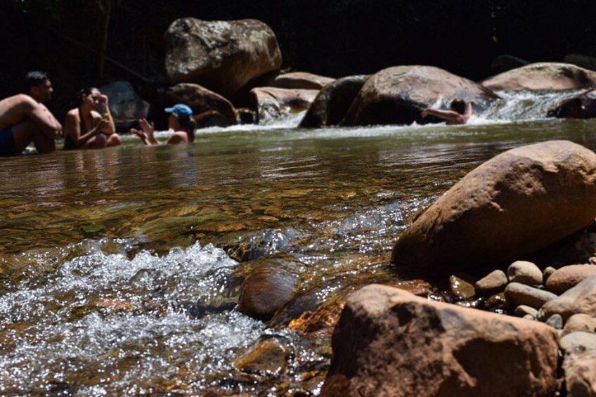 A clear stream with rocks, people sitting and standing in the water, and a large rock in the foreground. 2