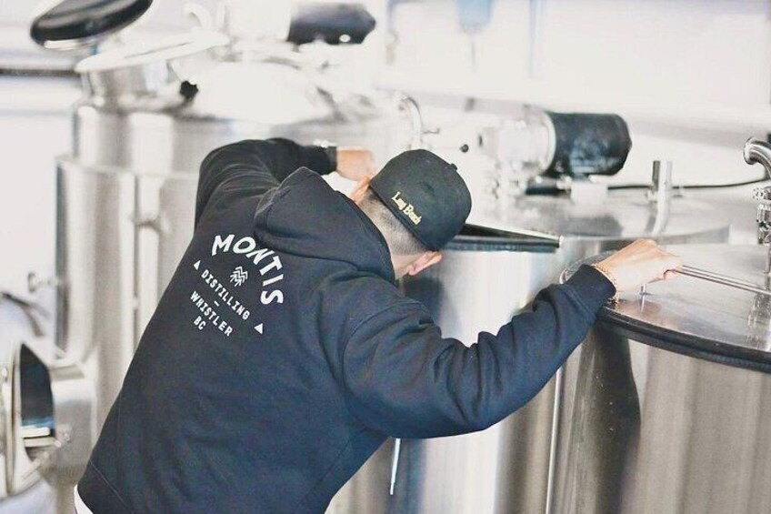 A person wearing a black hoodie with 'MONK'S' and 'A DISTILLERY WRESTLING' printed on the back, working with a large stainless steel container in a kitchen setting. 5