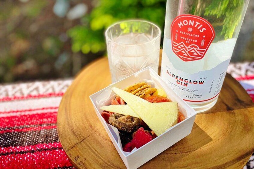 A bottle of Montis Alpine Glow Gin, a glass of water, and a box of assorted snacks on a wooden table outdoors. 3