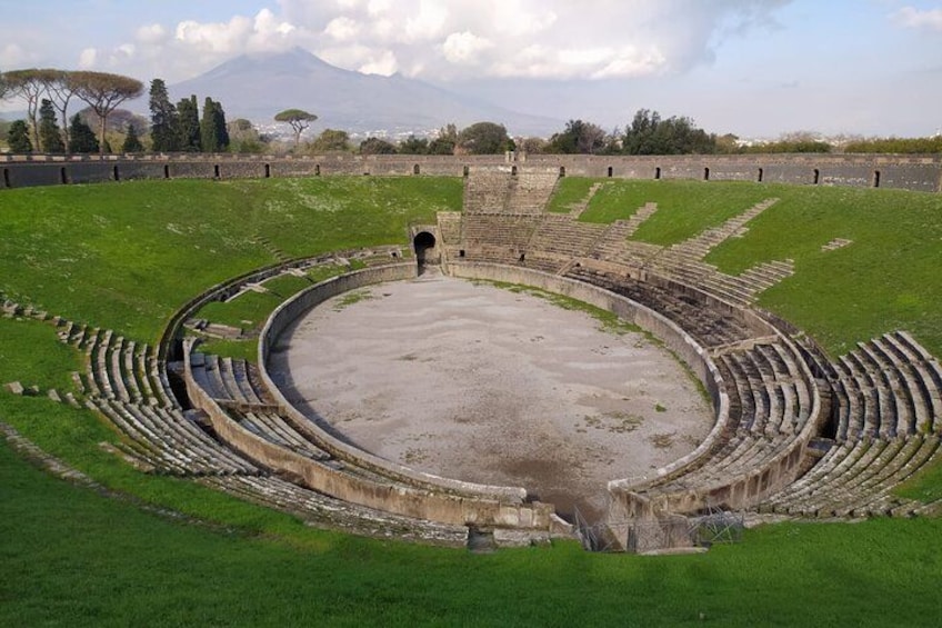 An ancient amphitheater with tiered seating and a central stage, surrounded by a grassy area and trees, with a mountain in the background. 2