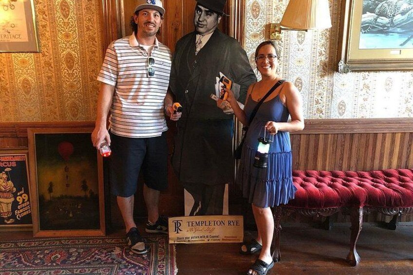 A man in a striped shirt and shorts, a woman in a blue dress, and a statue of a man in a suit. They are standing in a room with patterned wallpaper and a red cushioned bench. 5