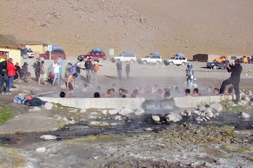 A group of people bathing in a natural hot spring surrounded by vehicles and a mountainous landscape. 3