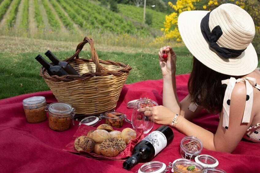 A picnic setup with a basket, jars, bread, wine, and a bottle on a red blanket in a grassy field. 5