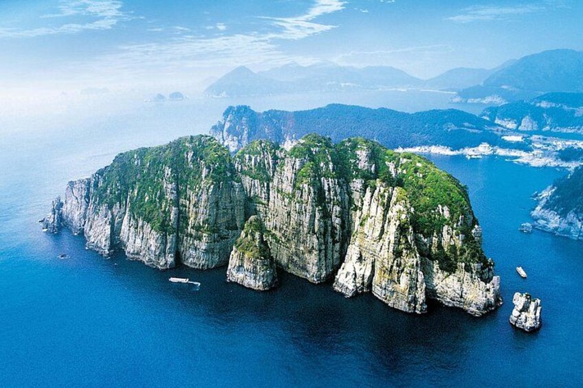 A coastal landscape with a prominent rocky island surrounded by clear blue waters, with boats visible in the distance. 4