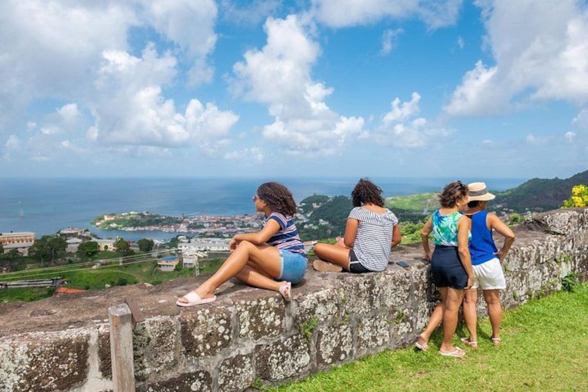 Quattro persone sedute e in piedi su un muro di pietra con vista su un paesaggio costiero. 1