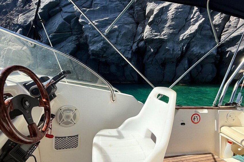 A boat's interior with a steering wheel, a white cushioned seat, and a rocky coastline in the background. 4