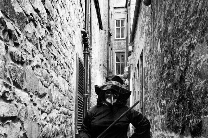 Un callejón estrecho con paredes de piedra tosca, una persona con sombrero y abrigo, y un edificio con ventanas al fondo. 2