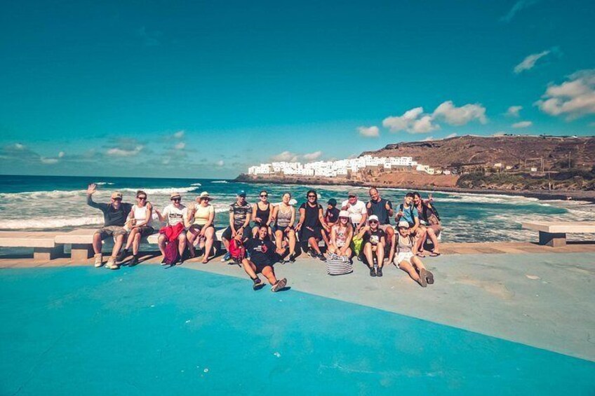 A group of people sitting on a concrete platform by the sea, with a coastal town in the background. 5