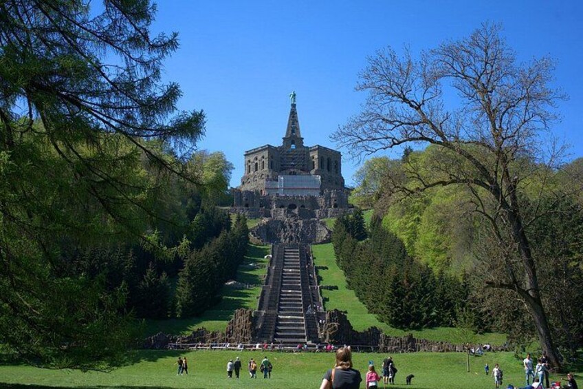 A castle with a steep staircase leading up to it, surrounded by trees and a grassy area with people. 2