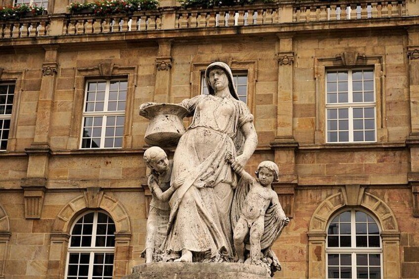A stone statue of a woman with two children on a pedestal in front of a building with arched windows. 5