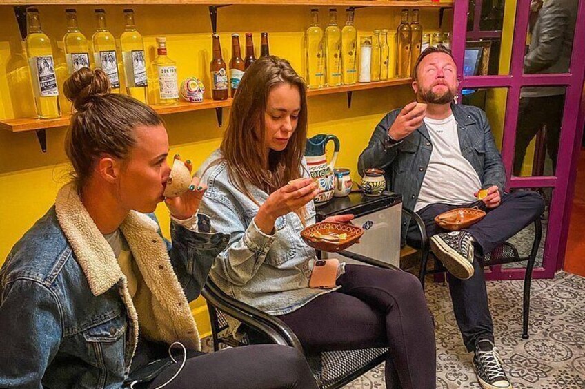 Three people sitting at a table with food, a shelf with bottles in the background. 4