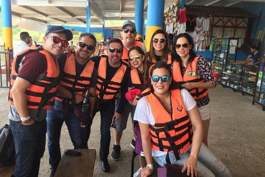 A group of people wearing orange life jackets, standing under a covered area with blue pillars. 2