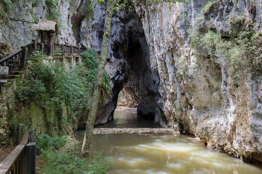 Entrada a una cueva con una escalera de madera que desciende, rodeada de abundante vegetación y un río caudaloso. 5