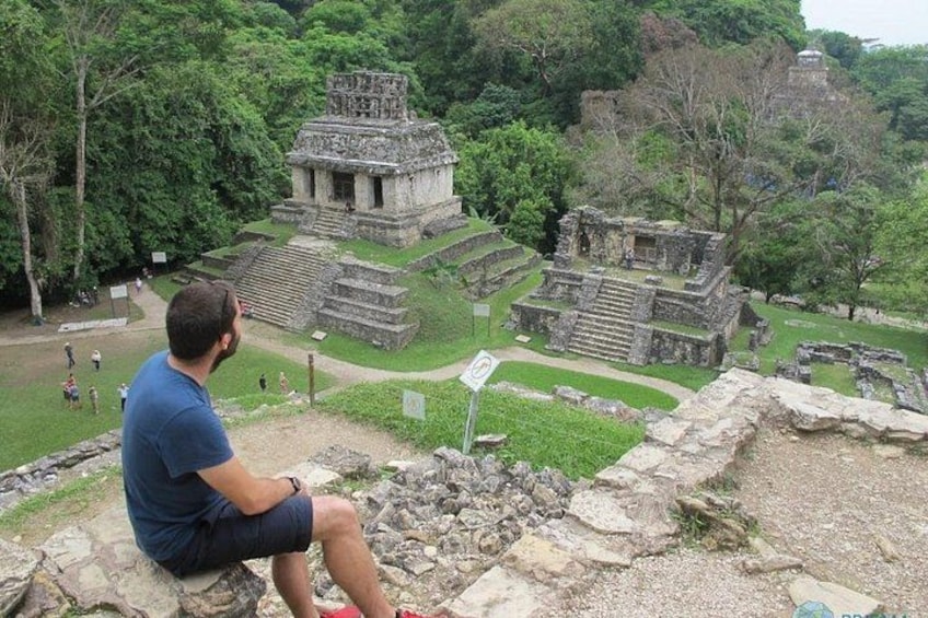 A man sitting on a stone ledge overlooking ancient Mayan ruins, surrounded by lush greenery. 2