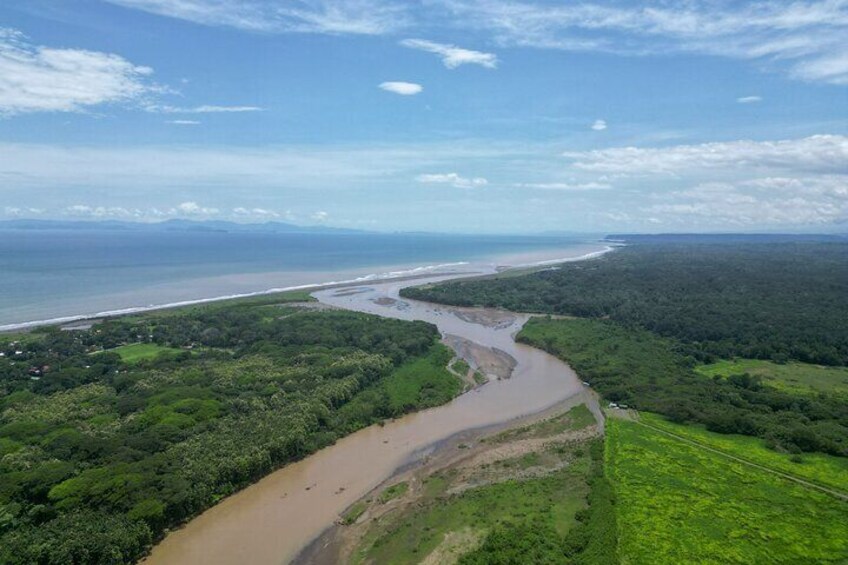 Ein Fluss schlängelt sich durch eine grüne Landschaft, im Hintergrund ein Strand und der Ozean. 4