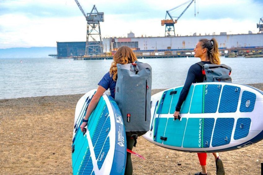 Dos personas sosteniendo tablas de surf en una playa con estructuras industriales al fondo. 3