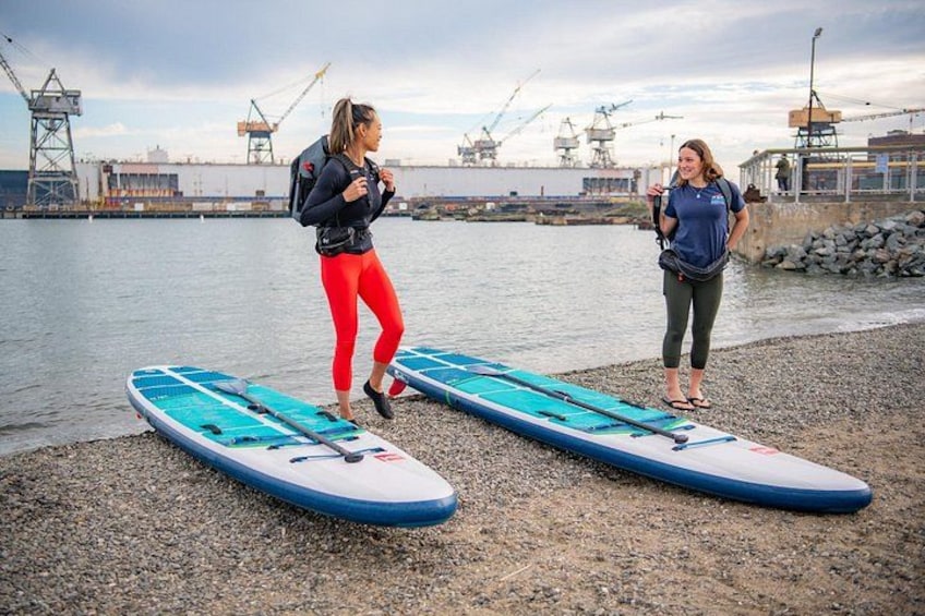 Två personer på en stenstrand med surfbrädor, ett vattendrag och industriella byggnader i bakgrunden. 2