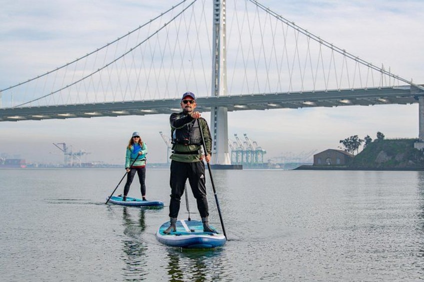 Dos personas haciendo paddleboard en un río tranquilo con un puente grande al fondo. 2