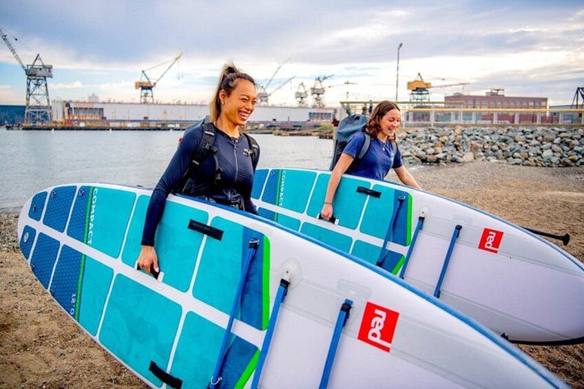 Dos personas sostienen tablas de surf en una playa con estructuras industriales al fondo. 1