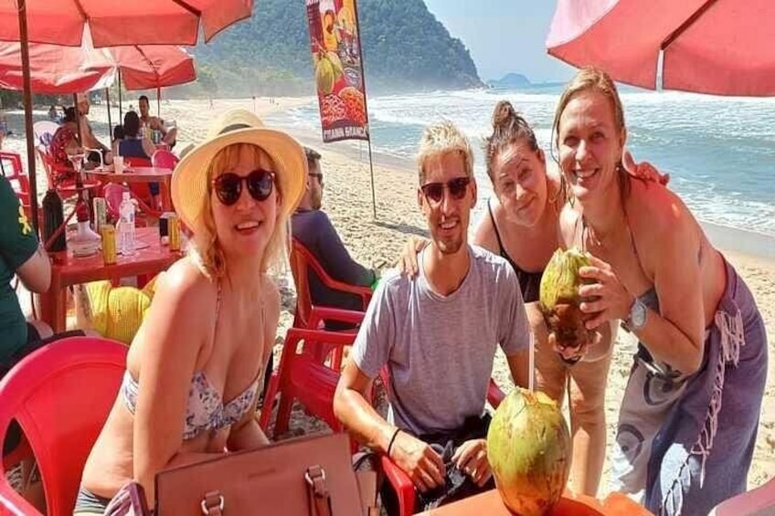En strandscene med mennesker siddende under parasoller, et bord med drikkevarer og en kokosnød i forgrunden. 1
