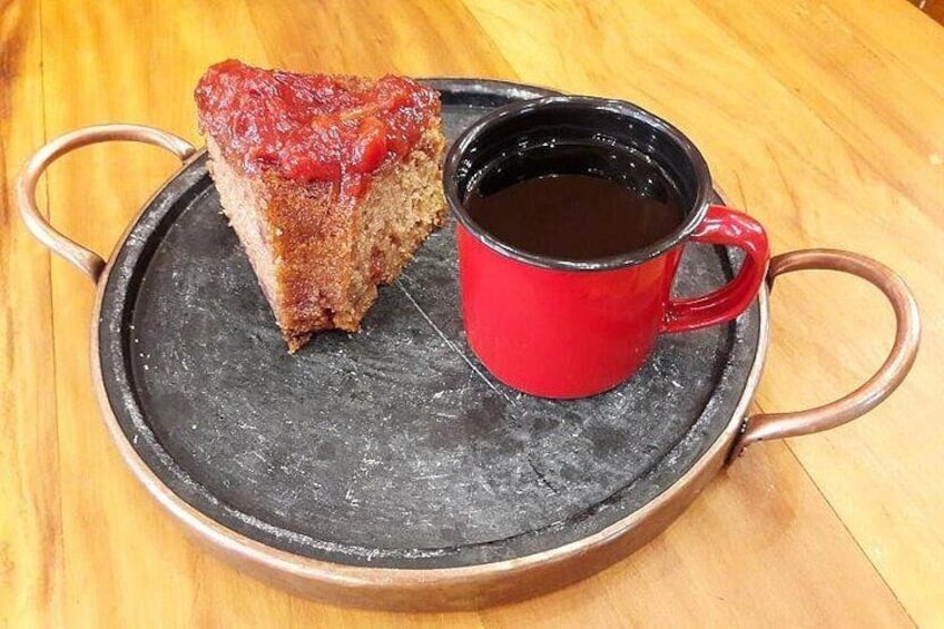 A slice of cake with a red topping on a black plate, accompanied by a red mug filled with a dark liquid, likely coffee, on a wooden table. 3