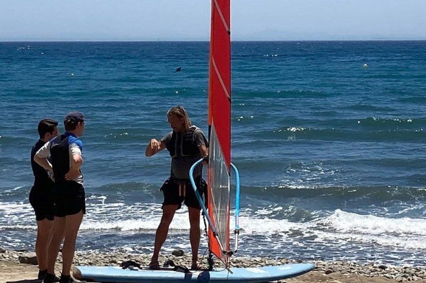 Eine Gruppe von Menschen am Strand, mit einem roten Segel, einem Surfbrett und dem Meer. 3