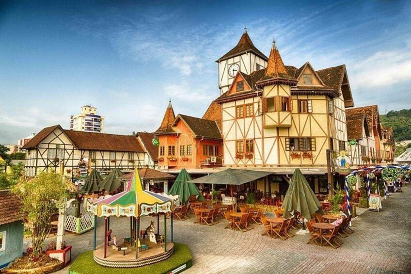 A traditional European town square with a prominent half-timbered building, outdoor seating areas, and a colorful carousel. 1