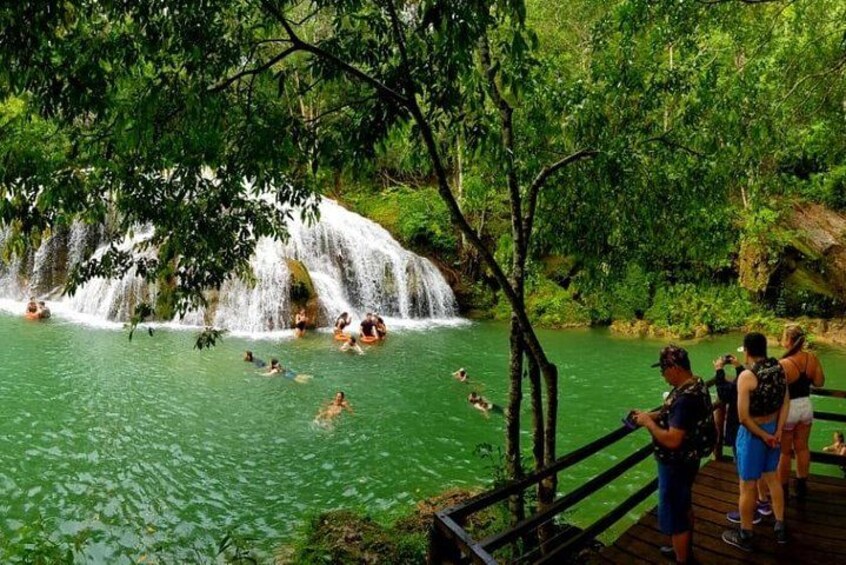 A waterfall in a forested area with people swimming and standing on a wooden deck. 3
