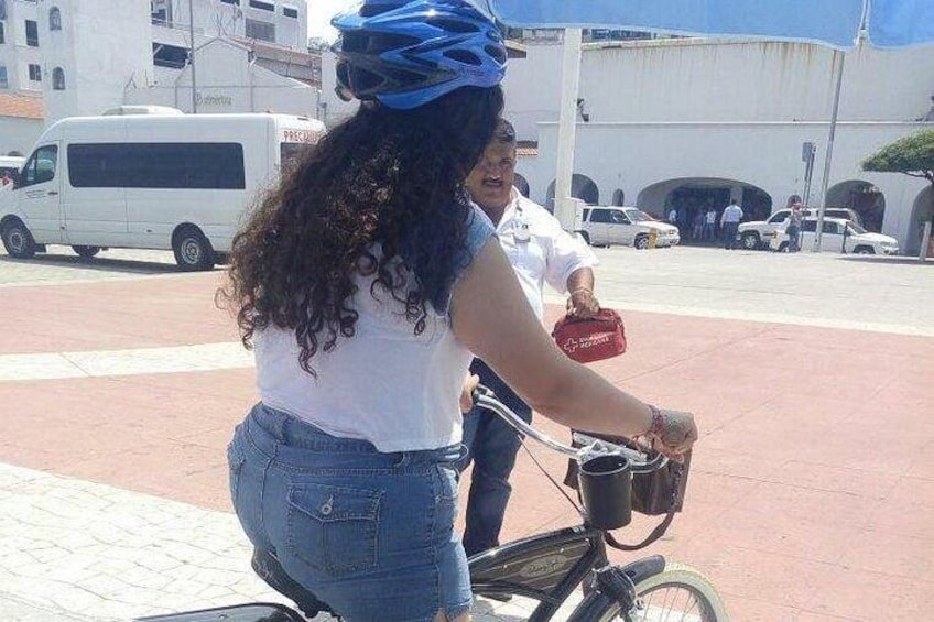 A person riding a bicycle on a paved road with a white van and buildings in the background. 3