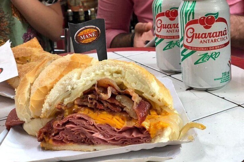 A sandwich with multiple layers of meat and cheese, a bottle of Guaraná Antarctica Zero, and a Mane sandwich stand sign. 5