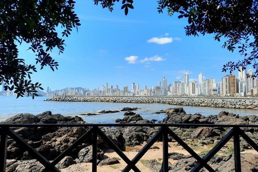 A coastal cityscape with a rocky shoreline, a clear blue sky, and a metal railing in the foreground. 1