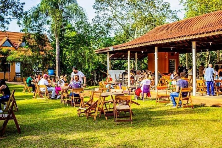 An outdoor dining area with wooden tables and chairs, surrounded by trees and a covered pavilion. 2