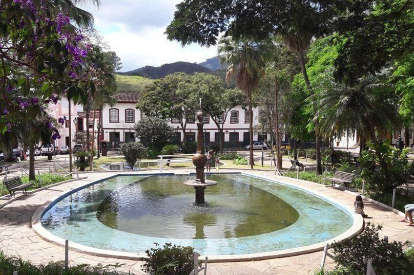 A circular fountain surrounded by trees and benches in a park with buildings in the background. 2