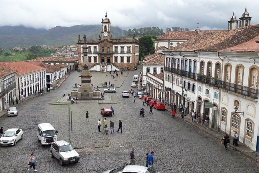 A cobblestone square with historic buildings, a central monument, and a clock tower. 3