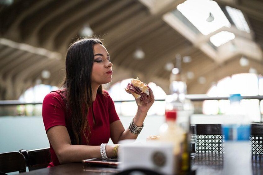 Una persona comiendo un sándwich en una mesa en un espacio interior amplio con techos altos y ventanas grandes. 1