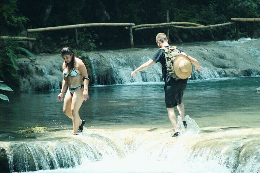 Una persona en traje de baño y otra con una mochila caminando sobre una roca cerca de una cascada. 1
