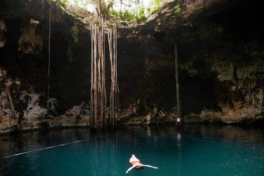 A clear blue pool with a person swimming, surrounded by rocky cave walls and hanging roots. 5