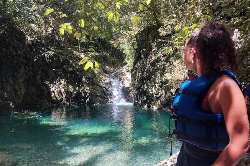 Una persona con chaleco salvavidas azul de pie junto a un cuerpo de agua azul y transparente, con una cascada en un área boscosa. 3