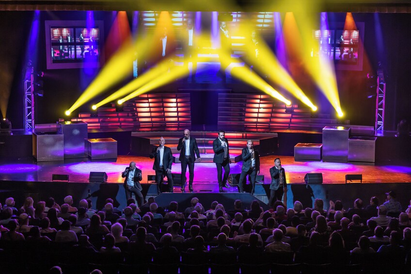 A stage with a group of people performing, a large audience, and stage lighting. 5