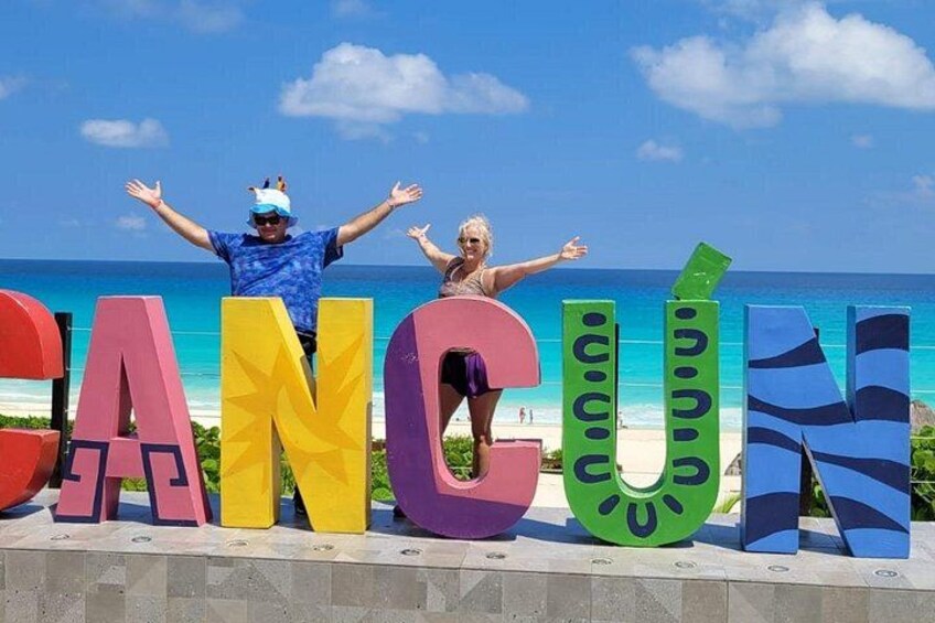 Una playa con letras grandes y coloridas que deletrean 'CANCUN' y dos personas de pie frente a ellas. 3