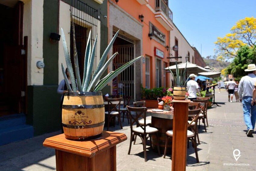 A street with outdoor seating, a wooden barrel planter with a plant, and a building with a sign. 2