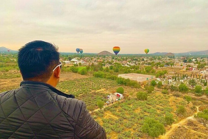Un hombre observando globos aerostáticos sobre un paisaje rural con campos y edificios dispersos. 2
