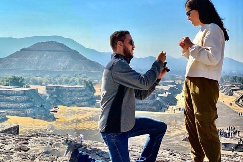 Una persona arrodillada proponiendo matrimonio a otra, con ruinas y montañas de fondo. 4