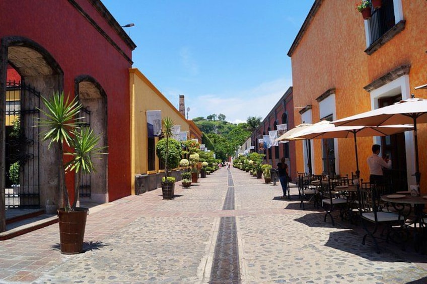 Una calle adoquinada bordeada de edificios, mesas al aire libre y macetas con plantas. 2