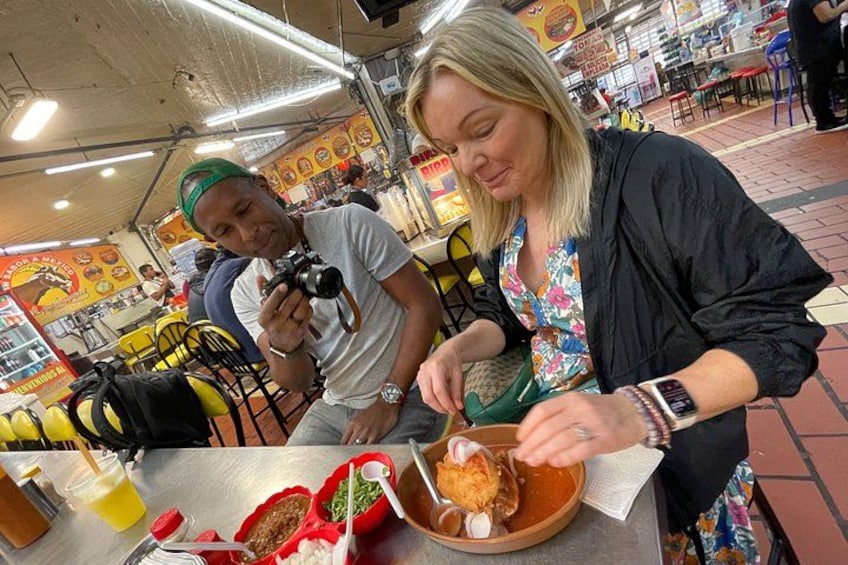 A person eating at a food stall with a camera and another person in the background. 5