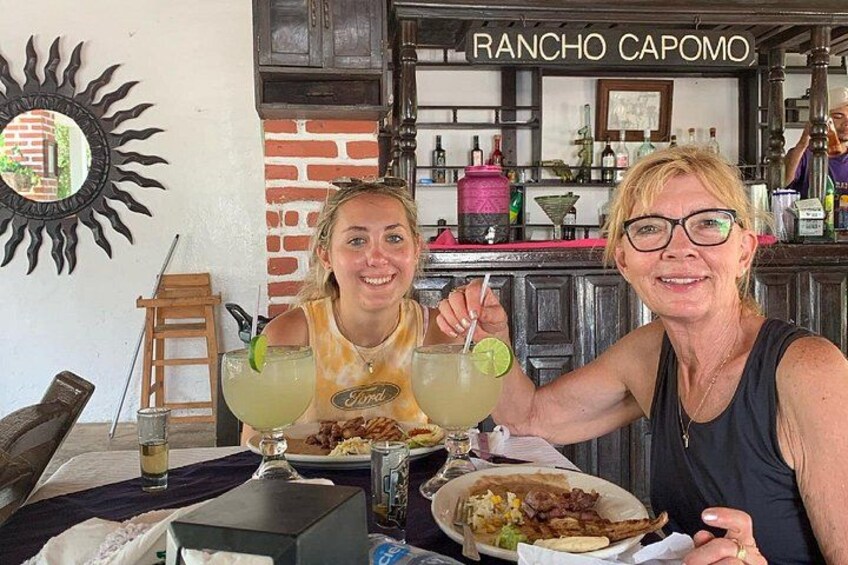 Two people at a table with drinks and food, in a restaurant with a brick wall and a sign reading 'RANCHO CAPOMO'. 3